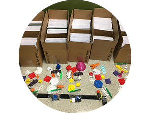 Five cardboard boxes filled with braille magazines and a smaller empty box. Colorful models are arranged of the boxes to be packed in the empty boces.